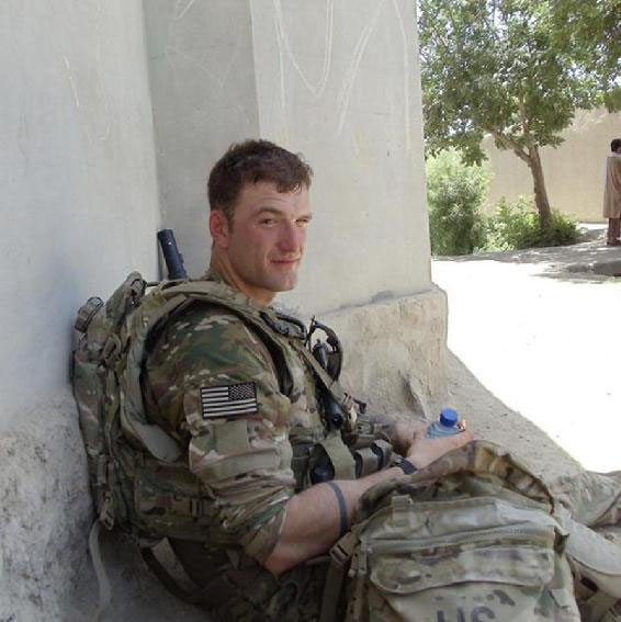 A photo of Joe Kassabian, dressed in green military fatigues and carrying a lot of gear, sitting in the shade of a building in what is presumably Kandahar in the time period of the book, looking sideways to the camera, holding a plastic water bottle.